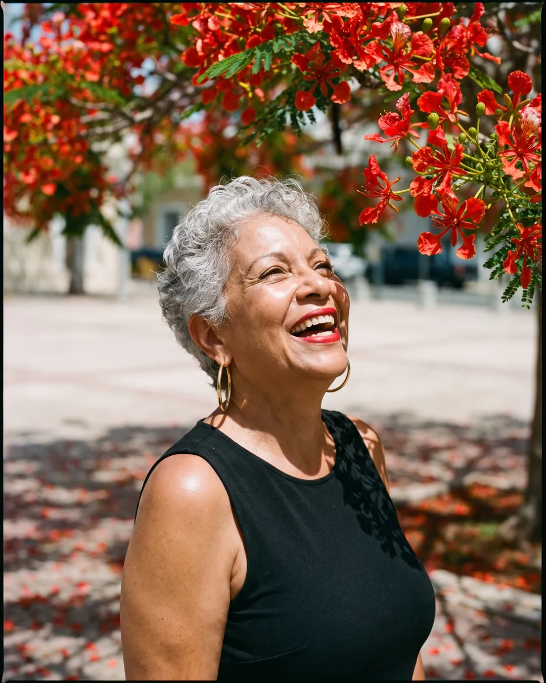 A woman with short silver hair laughs under a flamboyán tree in full red bloom