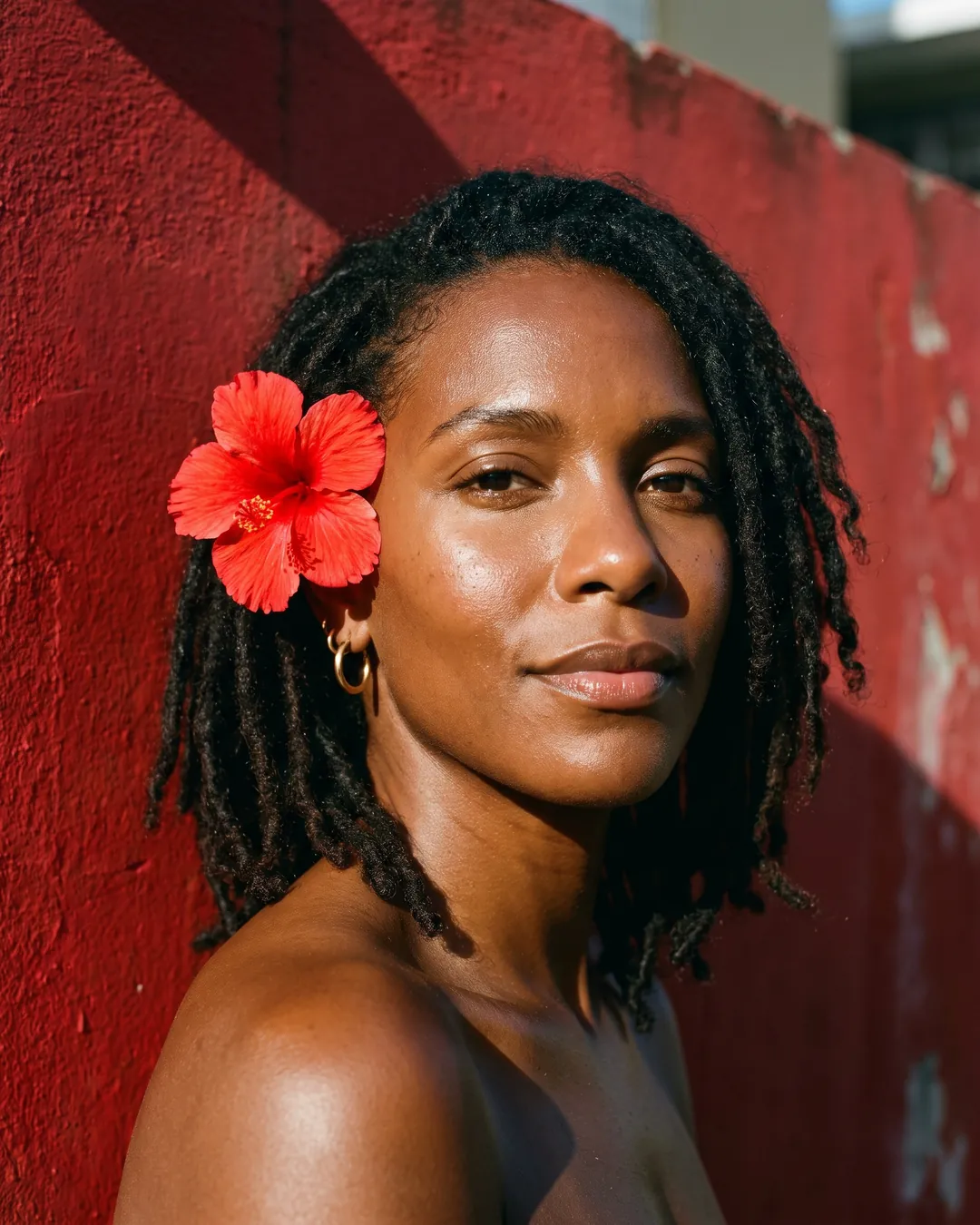 A woman with locs and a red hibiscus behind her ear looks into the camera against a weathered red wall