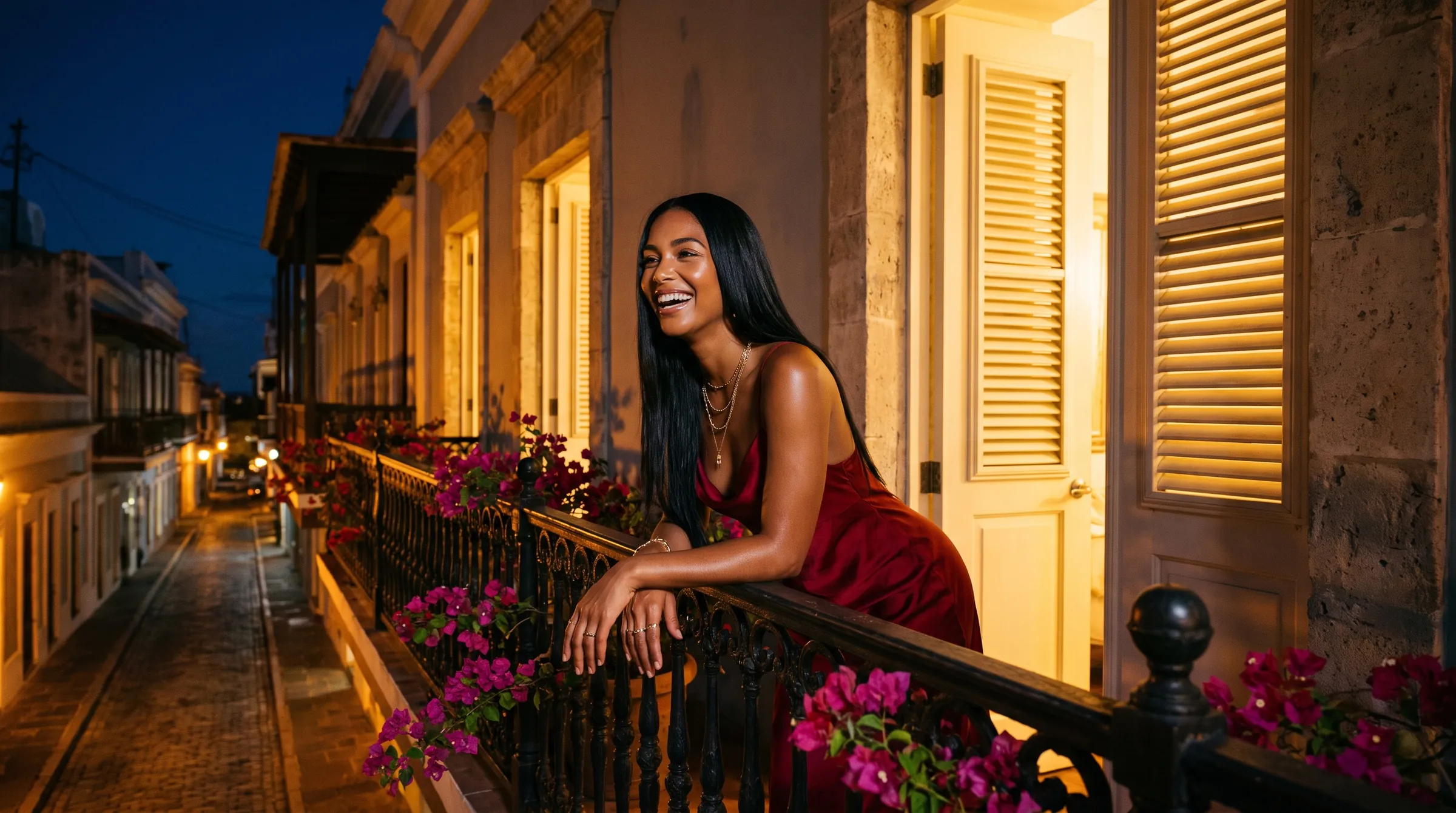 A woman in a red satin dress laughs on a wrought iron balcony over a lamplit street at night