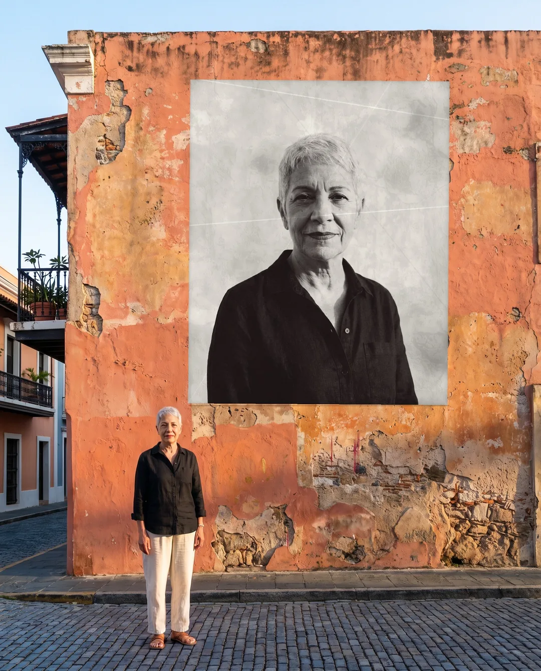 A two-story portrait of a silver-haired woman pasted on a coral wall in Old San Juan, the same woman standing on the cobblestones below it